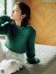 A woman sitting on a bed wearing a green sweater.